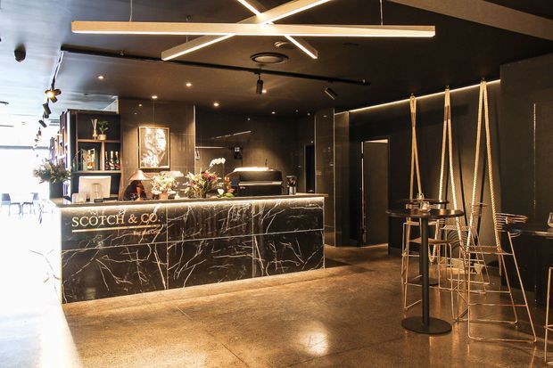 A dark, sophisticated commercial reception and bar area featuring a black marble-look reception counter with gold veining, illuminated lettering, black walls and ceiling, polished concrete flooring, decorative shelving, and distinctive suspended linear pendant lights. Rope-hung stools and small round standing tables line the right side, while a brighter seating area is visible beyond the reception zone.