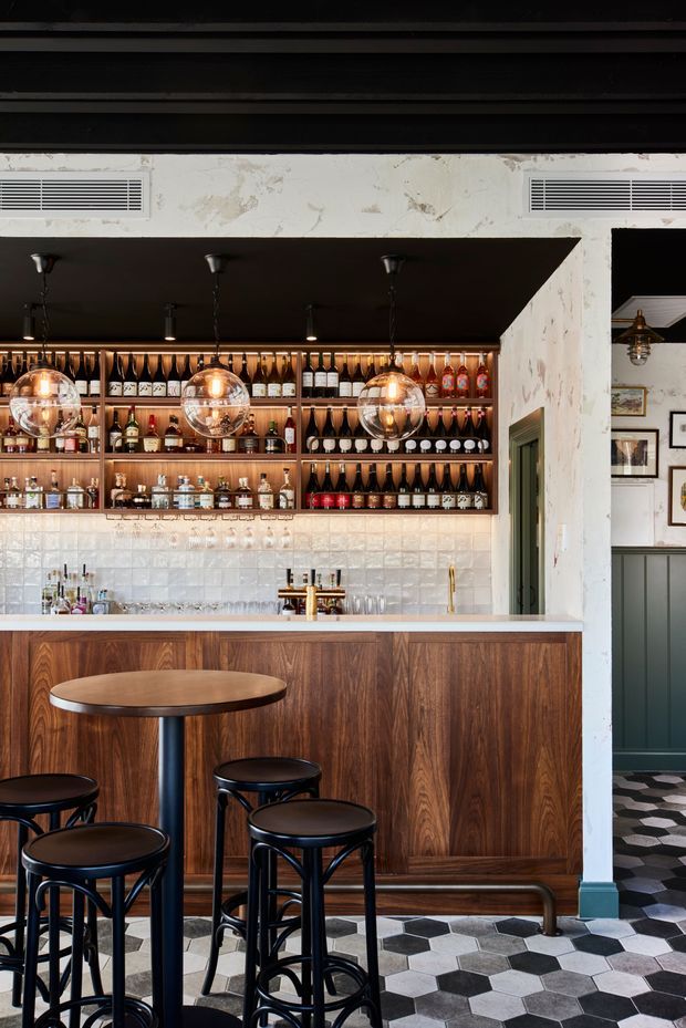 A moody, refined bar interior with a walnut-toned timber counter and back-bar cabinetry, stocked open shelving, white tiled splashback, brass fixtures, and three spherical pendant lights. Black stools and a small round standing table sit over a bold black, white, and grey geometric tiled floor. Dark ceiling beams, aged plaster, framed artwork, and a green-painted doorway add an eclectic hospitality character.