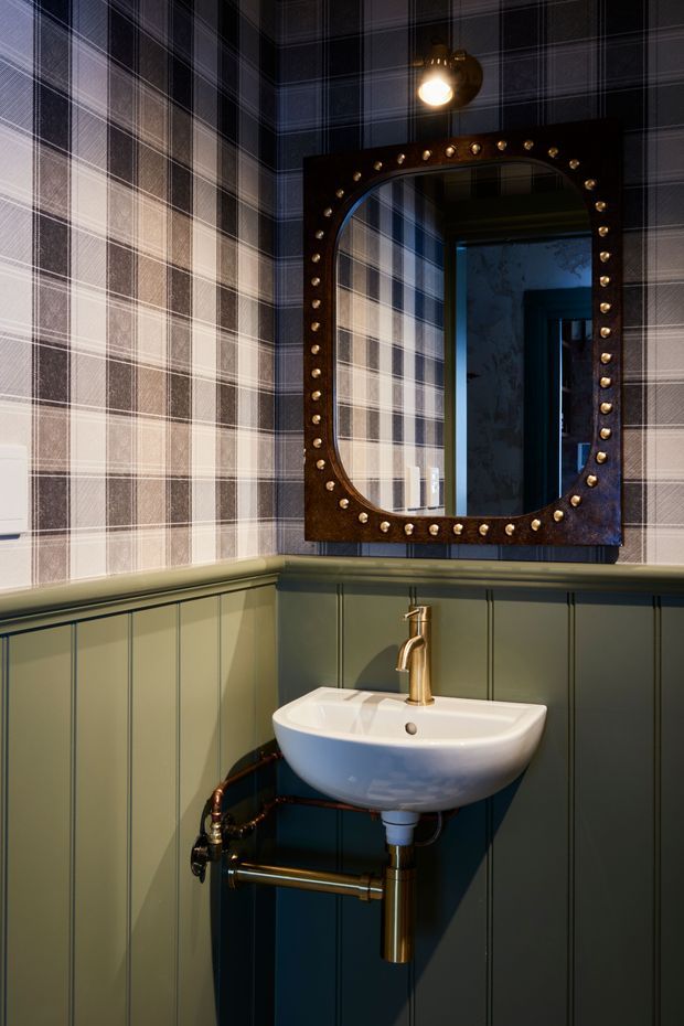 A compact, dramatically styled bathroom or powder room featuring a wall-mounted white basin with an exposed brass/gold-toned trap and plumbing, a brass tap, dark green vertical wall panelling, and dark plaid wallpaper above. A large rounded-rectangle mirror with an ornate dark frame and metallic stud detailing dominates the wall, illuminated by a small overhead fixture. The space has a moody, vintage-inspired and luxurious character.