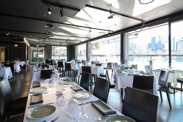 A contemporary waterfront restaurant dining room with numerous formally set tables, white tablecloths, dark angular chairs, and floor-to-ceiling glazing overlooking a city skyline and water. The space has dark polished flooring, a dark ceiling with exposed track lighting, and a refined, high-contrast hospitality interior.