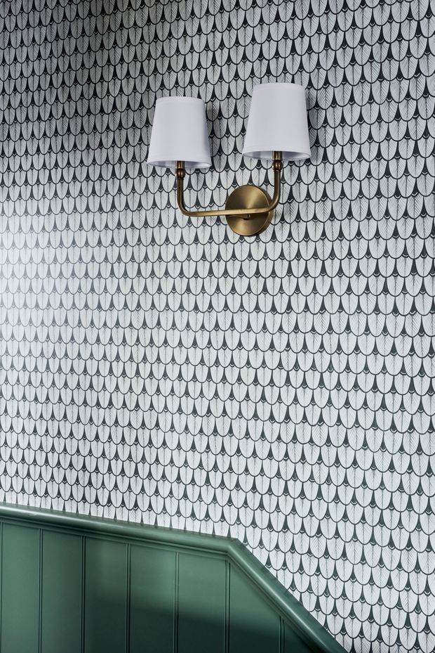 A close-up interior view of a stairway wall featuring scalloped patterned wallpaper in white and grey, a brass double wall sconce with white fabric shades, and dark green painted stair panelling and handrail at the lower edge. The composition is decorative and refined, with a strong repeating wall pattern and traditional detailing.