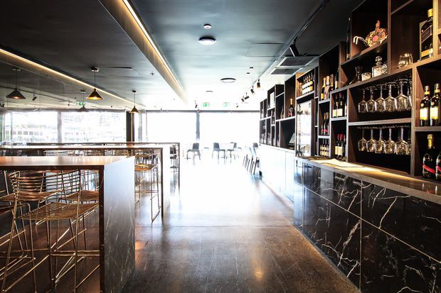 A contemporary commercial bar and dining space with a long black marble-faced bar, extensive dark timber display shelving, hanging wine glasses, and illuminated bottle storage. High tables with metal-framed stools occupy the left side, while bright full-height glazing opens the space toward the street. The palette is dark and refined with strong linear lighting and polished surfaces.