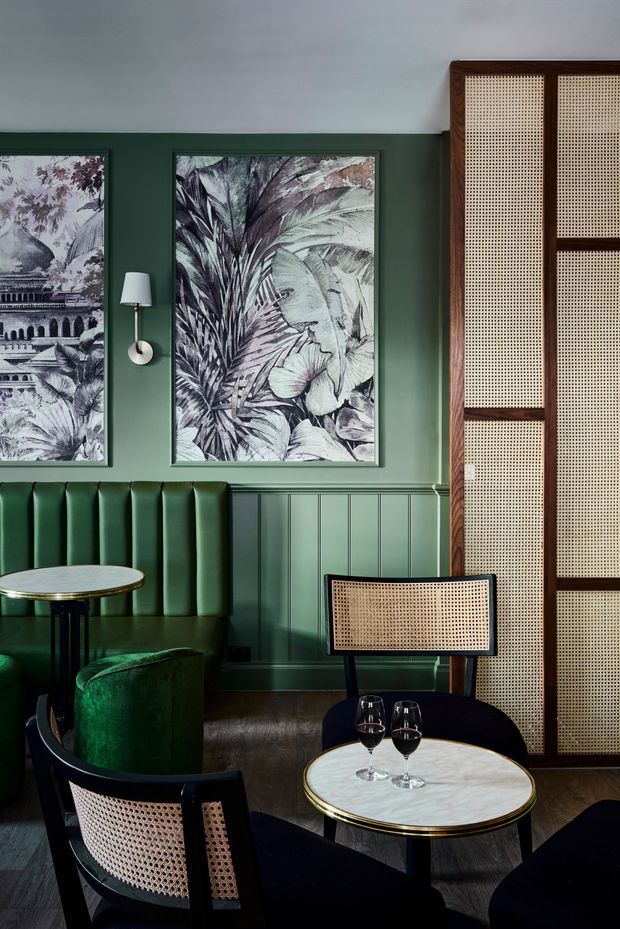 A richly styled hospitality lounge or bar interior with deep green walls, vertical green wainscoting, framed botanical artwork, and dark timber flooring. A tall timber-framed cane screen forms a feature wall on the right. The space contains green upholstered banquette seating, black-framed cane-back chairs, small round marble-topped tables with brass edging, wall sconces, and wine glasses. The overall atmosphere is intimate, refined, and moody.