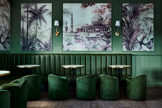 A richly styled dark green lounge or bar interior with a long channel-tufted banquette, compact round tables, and curved upholstered armchairs. The walls feature botanical and architectural artwork, brass wall sconces, and green vertical panelling. Dark timber-look flooring and deep emerald furnishings create a moody, refined atmosphere.