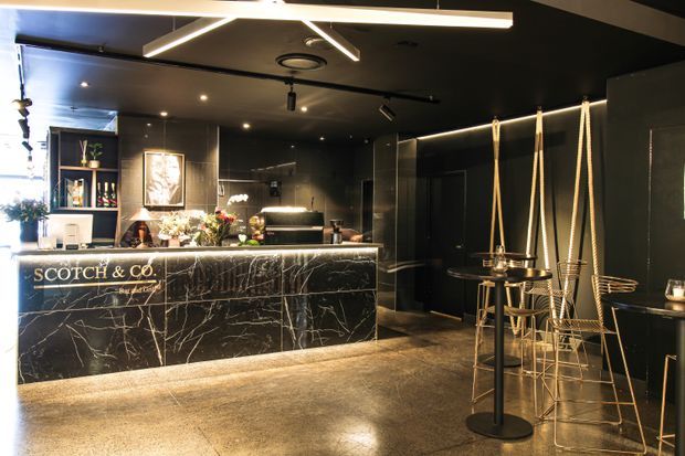 A dramatic, moody commercial bar or hospitality interior branded “Scotch & Co.” The space features a black marble-look bar counter with bright integrated lighting, dark walls and ceiling, illuminated display shelving, polished concrete flooring, geometric linear ceiling lights, and a small high-top seating area with metal stools and rope-like suspended details.