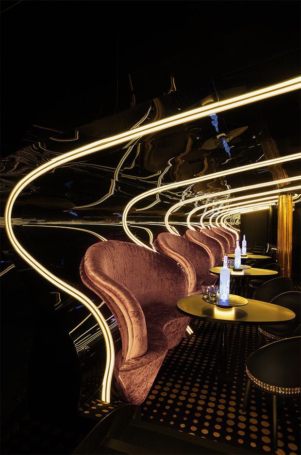 A dramatic, intimate hospitality interior with a row of sculptural blush-velvet wingback chairs arranged beside small round black-and-gold tables. Continuous glowing curved linear lighting is reflected across glossy black mirrored walls and ceiling, creating a tunnel-like effect. The floor has a black carpet with evenly spaced gold circular dots, and blue-lit table lamps provide small accent highlights. The space appears to be a sophisticated lounge, bar, or nightclub seating area.