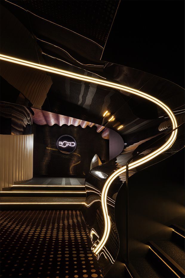 A dark, highly stylised commercial interior with a glossy black sculptural staircase or stepped circulation element, curved illuminated LED strips, reflective black surfaces, patterned carpeting, ribbed wall panels, and metallic wave-like ceiling and wall details. Warm golden lighting creates a dramatic futuristic atmosphere. A backlit circular sign is visible on the feature wall.