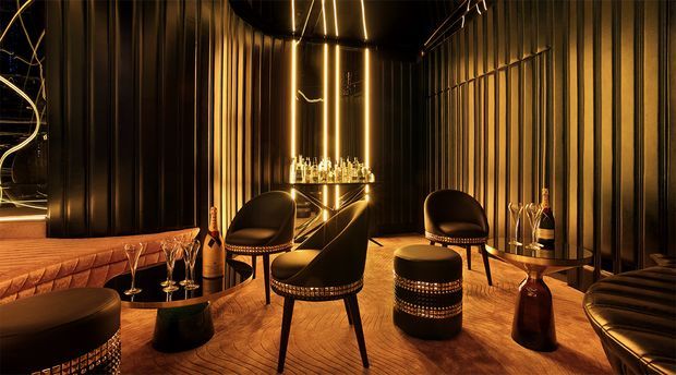 A dramatic, dark-toned home bar or lounge with black vertically fluted wall panelling, warm linear pendant lighting, herringbone timber flooring, and a compact illuminated bar counter. Curved black leather lounge chairs and stools surround low tables, with bottles and glassware displayed on the bar and side tables. The space has a luxurious, intimate, art-deco-inspired atmosphere.