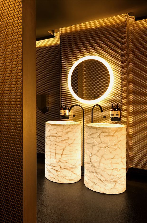 A dramatic, dimly lit bathroom featuring two sculptural freestanding vessel basins with pale textured surfaces, matte black arched tapware, and a large circular illuminated mirror. The walls and ceiling are clad in a repeating bronze-gold perforated metal pattern, with integrated warm LED lighting and a dark floor. The overall design is bold, luxurious, and highly atmospheric.