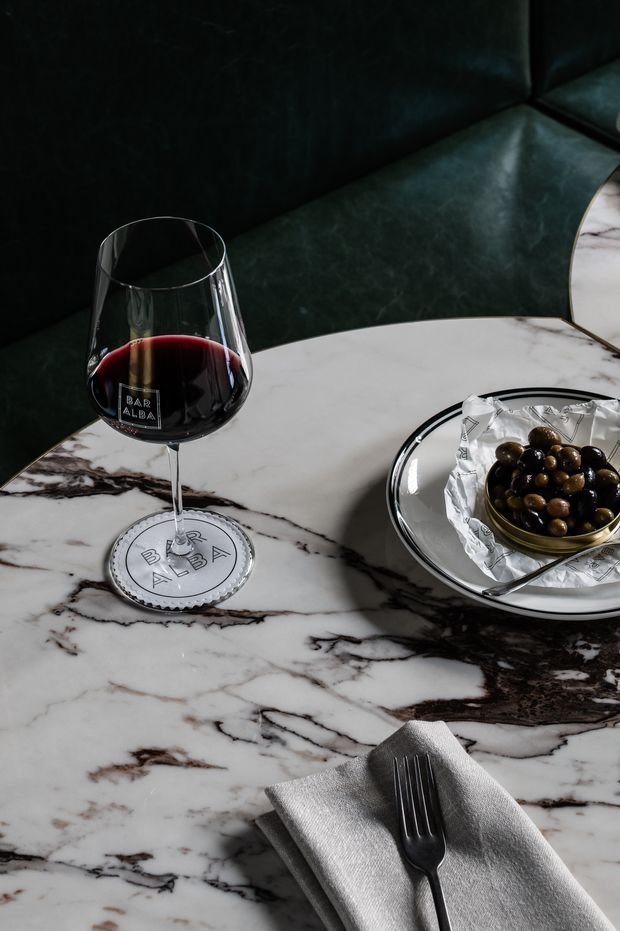 Close-up of a moody, refined dining setting featuring a dark green upholstered banquette, a white marble tabletop with dramatic dark veining, a red wine glass, olives on a reflective plate, and neatly folded grey napery with a fork.