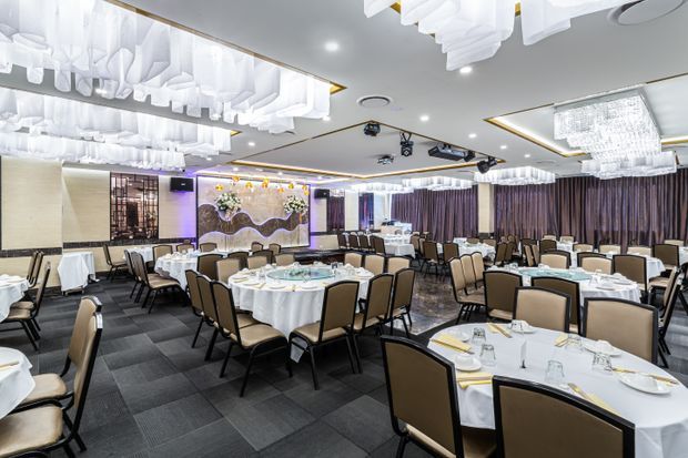 A large, formal banquet or restaurant dining room with numerous round tables dressed in white linens, upholstered dining chairs, dark patterned carpet, full-height curtains, decorative wall panels, and elaborate suspended ceiling light installations. The space appears suitable for events, weddings, or upscale dining, with a stage or decorative feature wall at the rear.