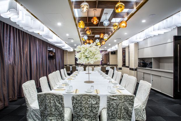 A long, formal dining room with a large white-clothed banquet table surrounded by upholstered dining chairs. The room features dark patterned curtains, dark flooring, pale wall panels, a decorative wood-paneled ceiling strip, and numerous amber and metallic pendant lights. A built-in sideboard and service area are visible along the right wall, with a floral centerpiece as the main table decoration.