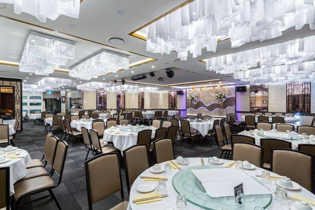 A large, brightly lit restaurant dining hall with numerous round tables dressed in white linens, upholstered dining chairs, place settings, and decorative floral displays. The ceiling features extensive sculptural white hanging installations, crystal-like chandeliers, recessed lighting, and gold trim. Mirrored wall panels, dark carpet flooring, and illuminated decorative screens contribute to an upscale banquet-restaurant atmosphere.