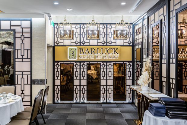 A refined seafood restaurant entry and dining area featuring an ornate geometric black-and-white partition and mirrored wall treatment, brass-toned Bar Luck Seafood Restaurant signage, multiple decorative chandeliers, dark tiled flooring, and elegantly set dining tables. The space has a dramatic, upscale hospitality interior with Art Deco-inspired detailing.
