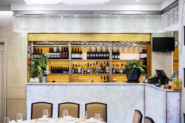 A refined bar counter in a restaurant or hospitality space, featuring a veined light stone-clad front and side, illuminated shelving stocked with wine and spirits, hanging glassware, potted plants, and a large crystal-like rectangular pendant light. The palette is light and warm with brass-gold shelving accents and dark timber or black details.