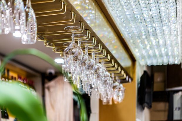 Close-up interior view of a sophisticated bar area, featuring rows of inverted wine and cocktail glasses suspended from brass-finished overhead racks. A large crystal-like decorative light fixture and reflective ceiling details create a luxurious, glamorous atmosphere, with dark timber and warm-toned finishes in the background.