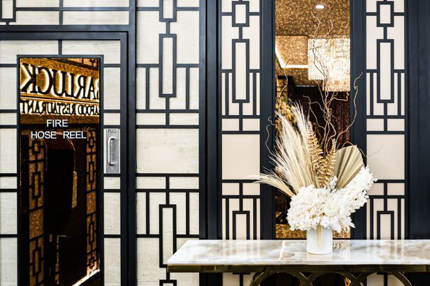 A refined interior entryway featuring a black-and-white geometric decorative screen and a dark glass fire hose reel cabinet at left. A slim marble-topped console table with an arched dark metal base sits in front of the screen, styled with a white vase of dried grasses and white floral arrangements. Warm textured golden wall surfaces are visible through the screen openings.
