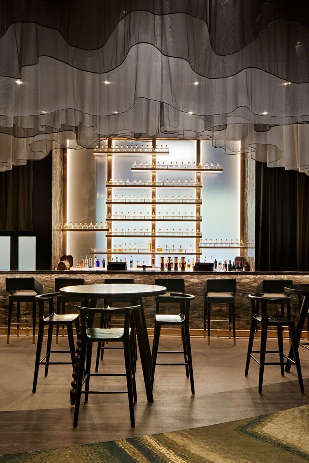 A sophisticated bar interior with a long stone-clad counter, illuminated backbar shelving displaying bottles and glassware, and black timber-framed bar stools and round tables. Dramatic layered translucent wave-like ceiling drapery, dark curtains, warm integrated lighting, and polished flooring create a moody, luxurious atmosphere.