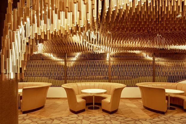 An ornate, warmly lit lounge or hospitality interior with curved tan leather armchairs arranged around small round tables. The space features a highly distinctive sculptural ceiling installation made from numerous suspended cylindrical wood-toned elements, mirrored or reflective surfaces, patterned screens, and a geometric stone or terrazzo-like floor.