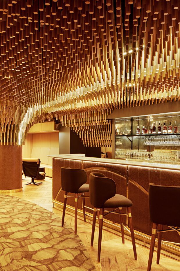 A richly detailed, warm-toned interior bar with a curved dark wood or bronze-finished counter, upholstered bar stools, illuminated bottle and glass shelving, and a dramatic suspended ceiling installation made from hundreds of hanging cylindrical wood-toned elements. The floor combines patterned carpet with light herringbone timber, while curved walls and integrated lighting create an intimate, luxurious atmosphere.