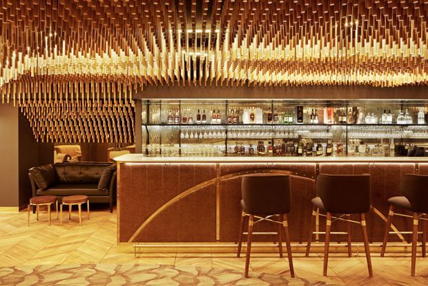 A richly detailed luxury home bar with a long curved-front bar counter, upholstered bar stools, illuminated glass shelving displaying bottles and glassware, and a lounge seating area. The room is defined by an elaborate suspended timber ceiling installation and matching timber wall treatment, with warm amber lighting and patterned timber flooring.