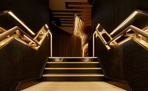 A dramatic, dimly lit stairway with dark textured wall finishes, black stair treads, warm integrated LED strip lighting, and angular metallic handrails. Timber-toned flooring and ceiling slats add warmth, while a reflective or glazed section and illuminated linear feature elements extend the view into the upper landing.