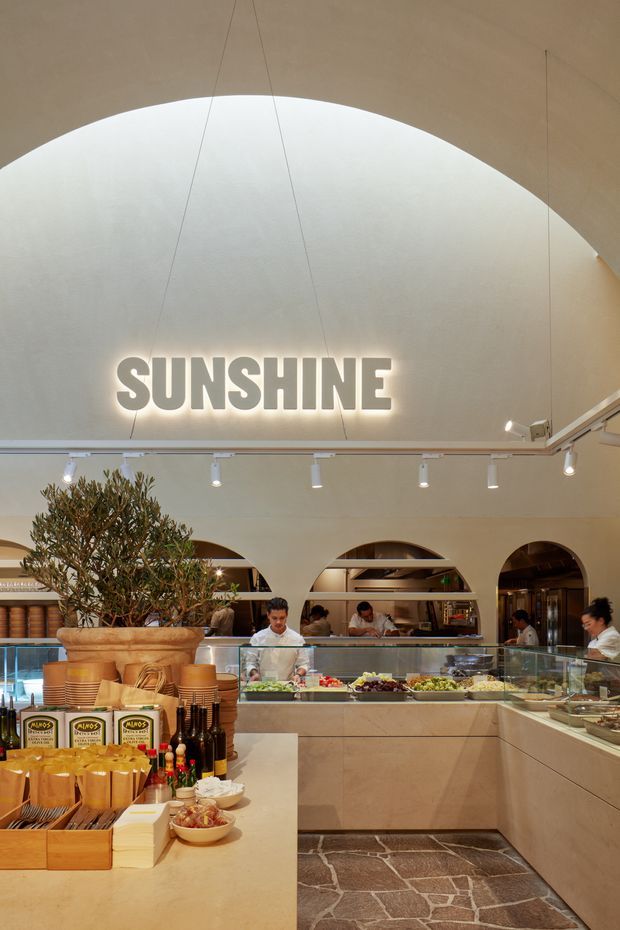 Interior view of a contemporary food-service venue or café with a large curved, vaulted ceiling and illuminated “SUNSHINE” signage. A central glass display counter presents prepared food, with staff working behind it and shelving visible through repeated arched openings. The space uses warm beige finishes, integrated track lighting, timber-toned counters and shelving, and an irregular natural-stone floor section. A large potted tree and packaged food products occupy the foreground.