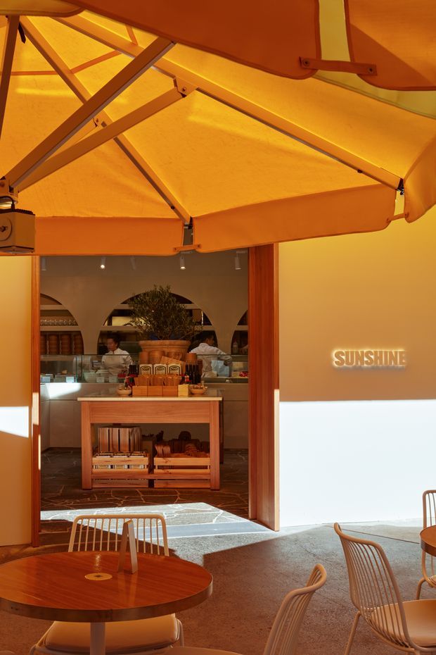 Warmly lit café interior viewed from a covered outdoor dining area, with a large orange retractable awning overhead, timber-framed entrance, service counter, display shelving, arched interior openings, and round timber dining tables with woven chairs. The space has a strong amber, earthy palette and a contemporary hospitality fit-out.