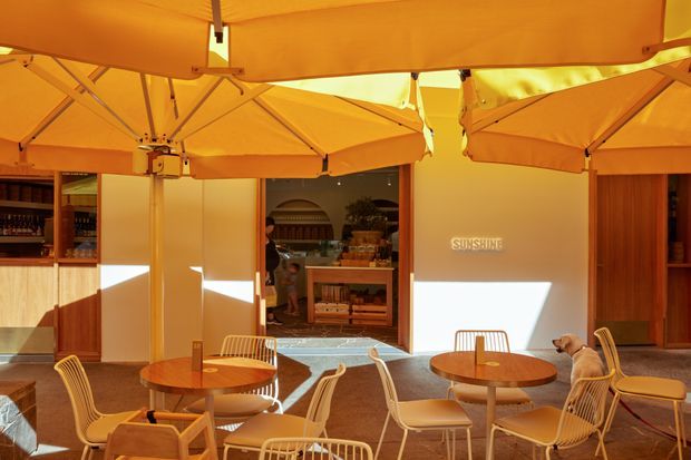 A warm, sunlit outdoor café or hospitality terrace beneath multiple large orange cantilever umbrellas. The space features round timber tables, woven and metal café chairs, textured paving, timber-framed service openings, built-in shelving, and a central counter area. Strong sunlight creates a golden, relaxed atmosphere, with the word “SUNSHINE” visible on the rear wall.