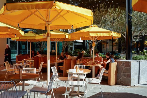 Outdoor dining terrace with numerous orange and yellow umbrellas shading round tables and chairs. The setting appears to be a contemporary hospitality venue or restaurant courtyard, with planted areas, glazed building façades, stone paving, and a low stone planter wall. The scene is lively and sunlit, with warm orange furnishings and umbrellas creating a strong visual identity.
