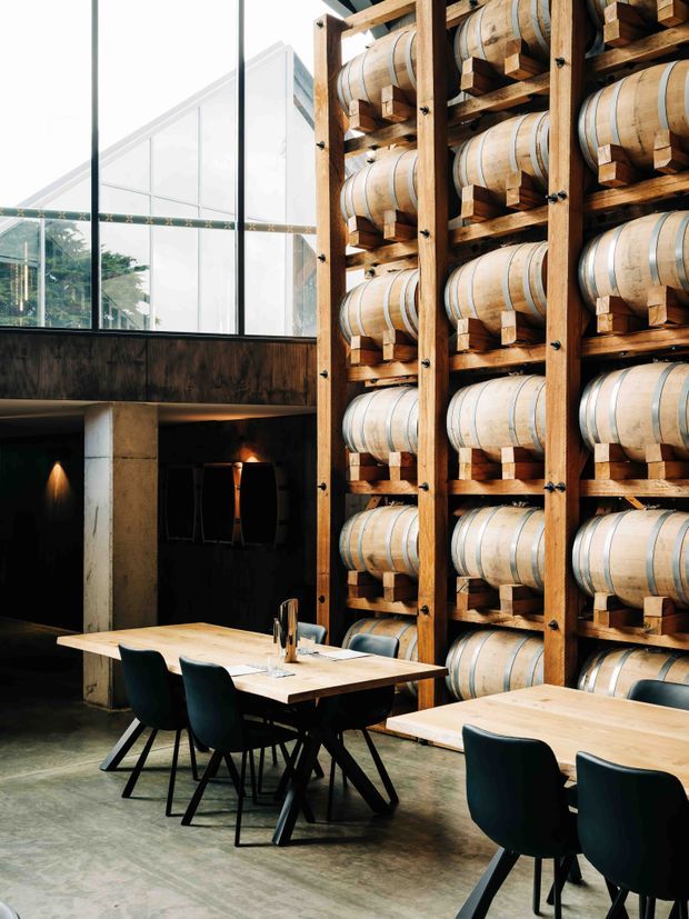A dramatic contemporary dining space with two light timber dining tables and dark upholstered chairs, set against a tall, full-height timber rack storing rows of large wooden barrels. The room has an industrial character with exposed concrete columns, a polished concrete floor, dark metal-framed glazing, and a high glazed roof or clerestory admitting abundant daylight. Warm pendant-like wall lighting contrasts with the dark lower-level background.