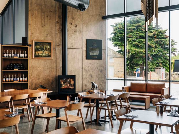 A stylish double-height restaurant or wine bar interior with extensive floor-to-ceiling glazing, warm timber wall panelling, exposed black ductwork, and a black freestanding fireplace. The space is furnished with numerous timber dining tables and chairs, leather banquette seating, and built-in bottle display shelving. Large windows overlook greenery and neighbouring buildings, while pendant lighting and a warm neutral palette create an inviting, refined atmosphere.