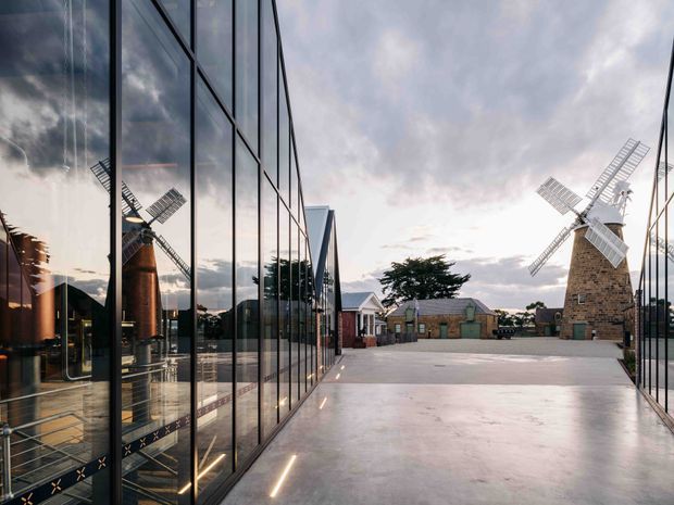 A dramatic architectural exterior view of a renovated industrial or heritage site at dusk. A broad concrete-paved forecourt is framed by tall, highly reflective glazed facades on both sides, mirroring the surrounding windmills and cloudy sky. A historic stone windmill and a second windmill structure stand in the background, alongside low heritage-style buildings. Warm linear lighting is integrated near the base of the glazing, creating a refined contrast between the contemporary glass architecture and the historic masonry structures.