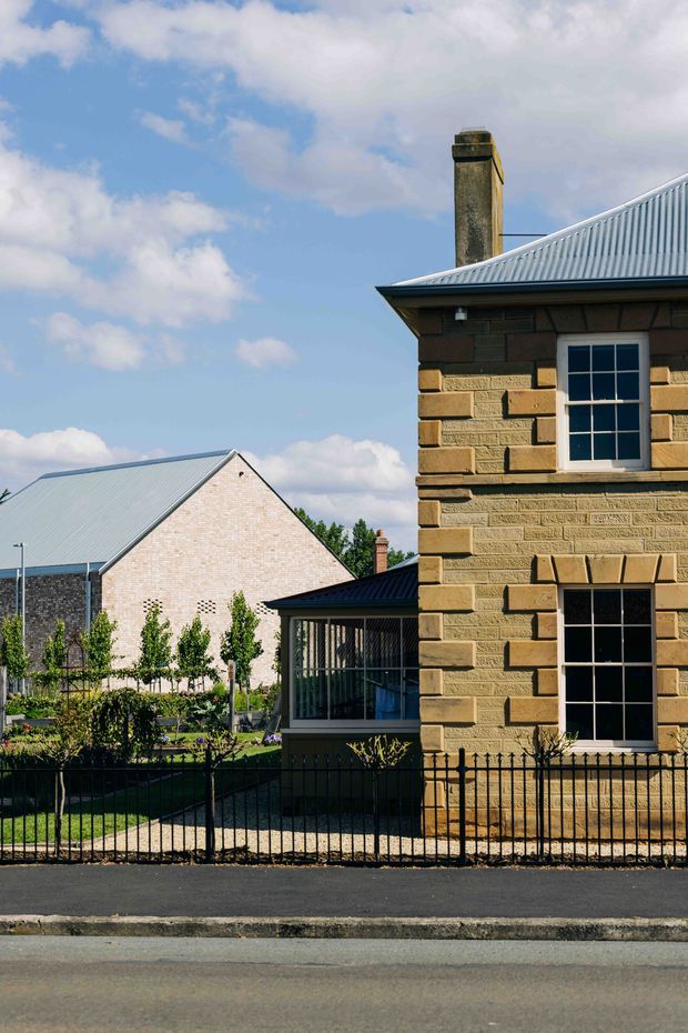 Exterior architectural photograph of a traditional masonry house, prominently showing a sandstone-clad facade with projecting quoins, vertically proportioned multi-pane sash-style windows, a corrugated metal pitched roof, and a tall masonry chimney. A landscaped front garden behind a black metal fence and adjacent brick buildings are also visible.
