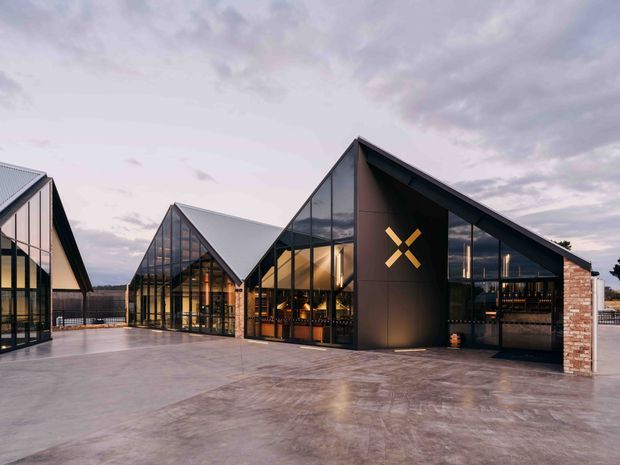 Exterior view of a contemporary commercial complex composed of several steeply pitched, angular gabled volumes. Extensive floor-to-ceiling glazing reveals warmly lit interior hospitality or retail spaces, while dark metal cladding, exposed brick end walls, and broad polished concrete paving create a refined industrial-modern character. A geometric illuminated logo is mounted on the central facade.