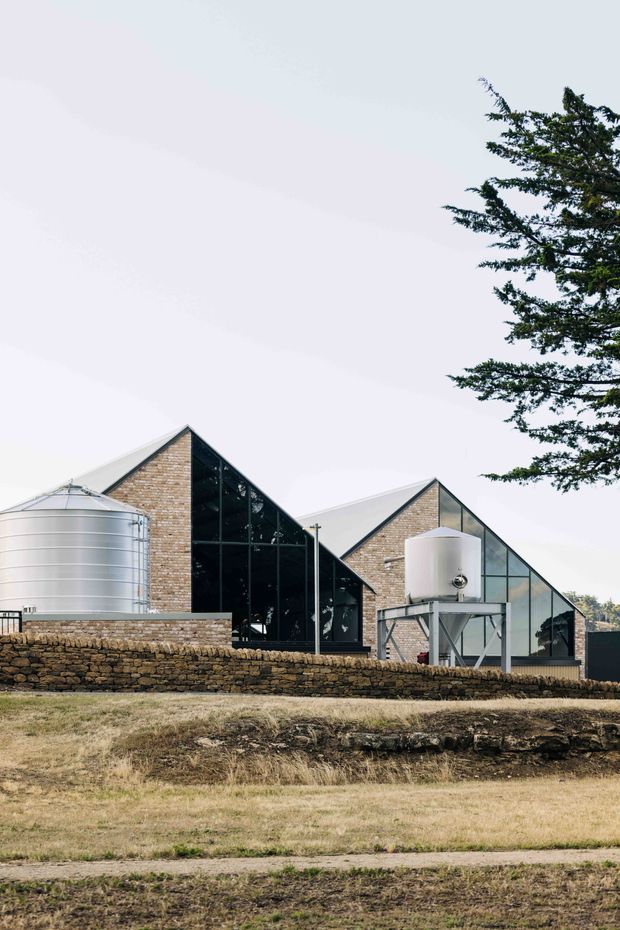 Exterior view of a contemporary rural or agricultural building composed of interconnected steep gabled volumes. The structures combine exposed tan brick gable walls with extensive black-framed glazing and pale metal roofs. Large cylindrical metal tanks and a stone retaining wall sit in front of the building, with dry grassland and a mature tree surrounding the site.