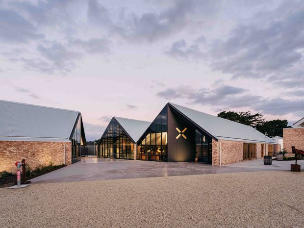 Exterior architectural photograph of a contemporary low-rise complex composed of several steeply pitched gabled volumes. The buildings combine pale grey metal roofing, dark framed glazed gable ends, black metal cladding on the central entry volume, and warm exposed brick walls. A broad gravel forecourt and paved approach lead toward the illuminated central entrance, photographed at dusk beneath a cloudy sky.