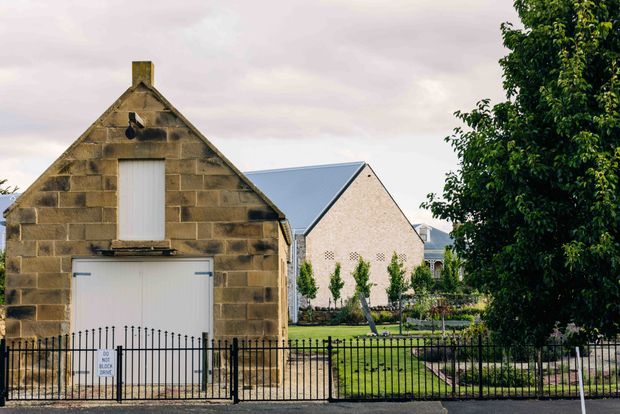 Exterior view of a small historic stone gabled building beside a larger pale masonry gabled building. The foreground structure has rough brown stone block walls, a steep pitched roof, a tall narrow upper opening with a white panel, and a large white garage-style door behind a black metal fence. Landscaped grounds and trees surround the buildings under an overcast sky.