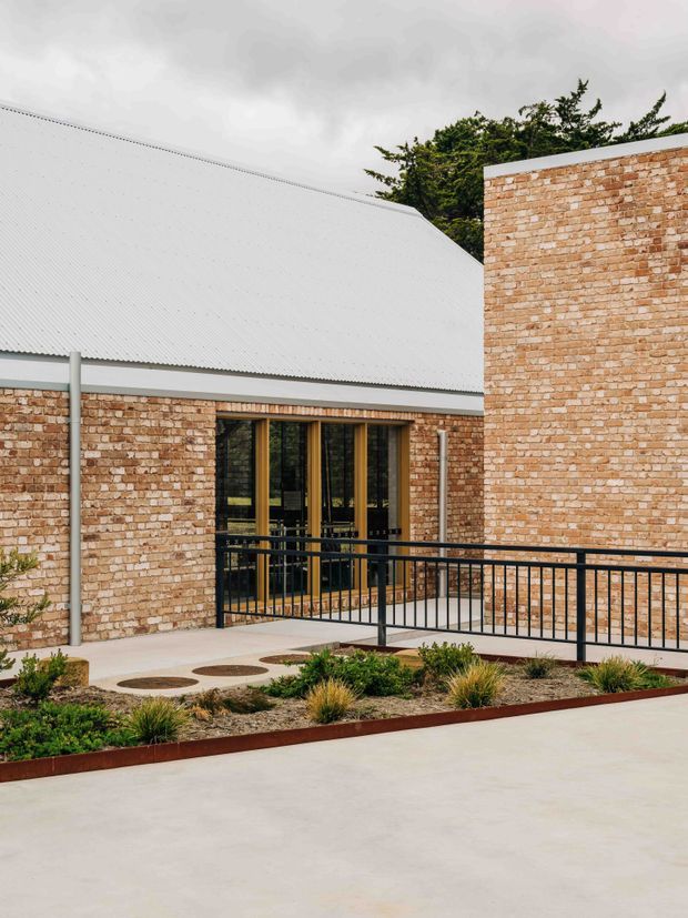 Exterior view of a low-rise brick building with a steep corrugated metal roof, tall glazed timber-framed entrance doors, and a black metal accessibility ramp railing. A landscaped garden bed and pale concrete paving occupy the foreground under an overcast sky.