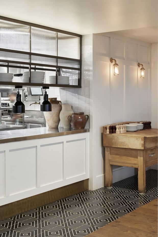 A refined restaurant or hospitality interior showing a service counter and open pass-through kitchen. White panelled cabinetry and walls are paired with dark metal-framed ribbed glass screens, white tiled splashback, black pendant lights, vintage-style wall sconces, stone counters, and a rustic timber side table. The floor combines a bold black-and-white geometric patterned surface with timber flooring.