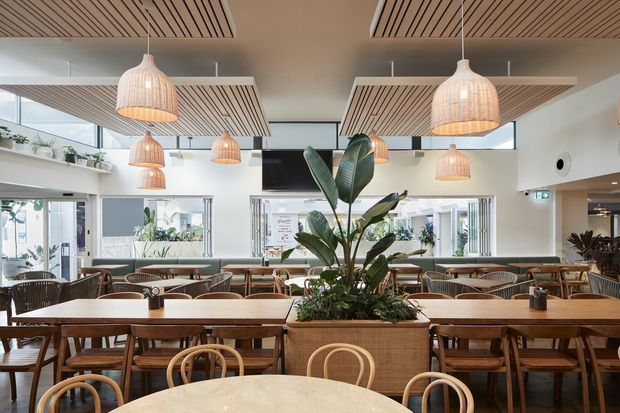 Bright contemporary hospitality dining space with extensive timber dining tables and bentwood chairs, a large central planter, woven pendant lights, and decorative timber slatted ceiling panels. The open layout includes broad glazing, indoor planting, and a refined natural-material palette.