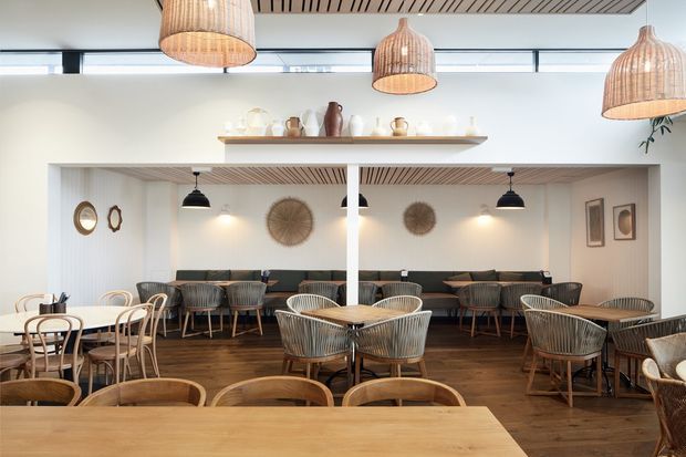 Warm, contemporary café interior with a broad timber communal table in the foreground, multiple timber dining tables and woven chairs, banquette seating along the rear wall, pendant lighting with woven shades, dark timber flooring, white walls, decorative shelving, and slatted timber ceiling detailing.