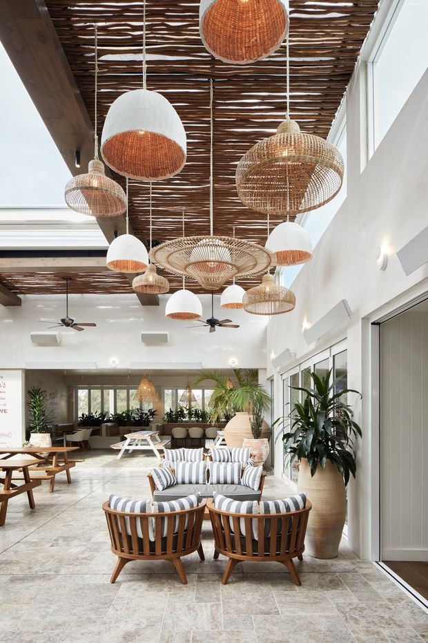 A bright, spacious hospitality-style lounge or lobby with high ceilings, exposed dark timber beams, a woven timber slat ceiling canopy, and numerous suspended woven pendant lights. The space features white walls, large glazed openings, pale stone-look tiled flooring, tropical plants, and timber lounge furniture upholstered in striped neutral fabric. Ceiling fans and abundant glazing reinforce the airy resort-like atmosphere.