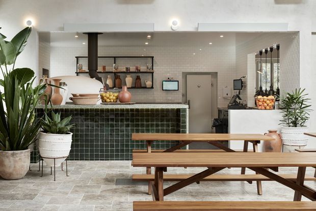 A stylish café or hospitality interior with a dark green tiled service counter, white tiled walls, open black metal shelving, pendant lighting, timber communal picnic-style tables and benches, and abundant indoor plants. The space has a warm contemporary aesthetic with natural timber and earthy decorative elements.