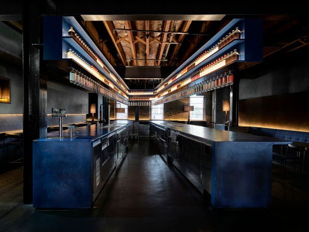 Dark, dramatic commercial bar interior with a long central aisle flanked by deep blue bar counters and extensive illuminated bottle shelving. The space features black walls, a glossy dark floor, exposed timber ceiling structure and services, industrial-style metal detailing, pendant and concealed warm lighting, and a rear brick feature wall with large windows.