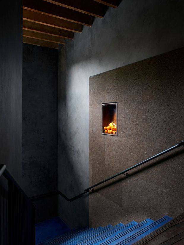 A dramatic, dimly lit stairway descending through a narrow, double-height space. Dark textured plaster walls and a warm brown feature wall are contrasted by exposed timber ceiling beams, a black metal handrail, and blue-toned stair treads. A small recessed wall niche displays oranges and provides a warm focal point.