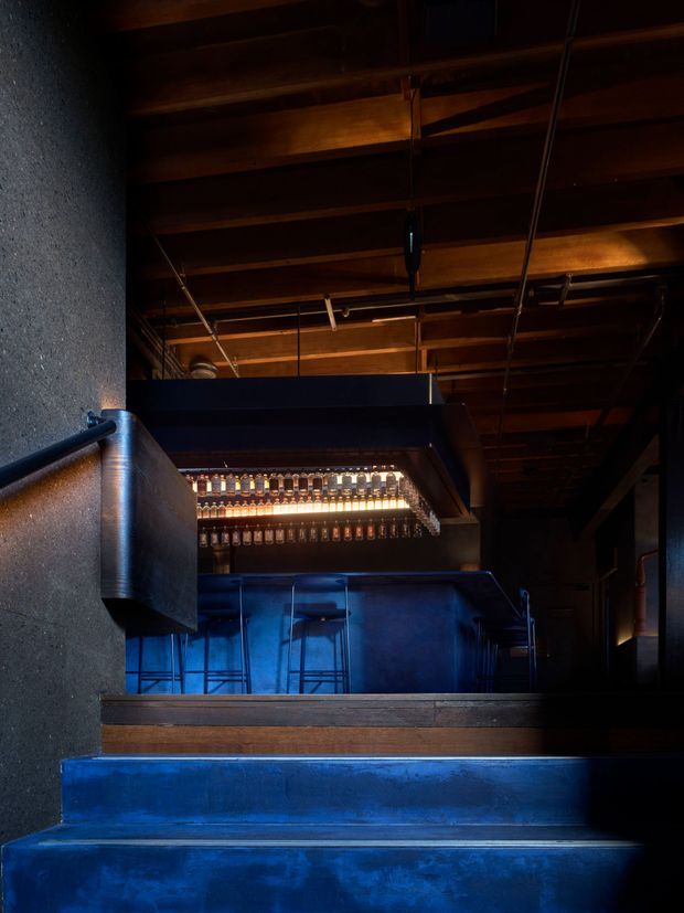 A dark, atmospheric bar or hospitality interior viewed from a short flight of blue-painted steps. The space features a deep blue bar counter and stools, a glowing backlit display of bottles or glassware, dark timber exposed ceiling beams, concrete walls, and dramatic blue-and-amber lighting.