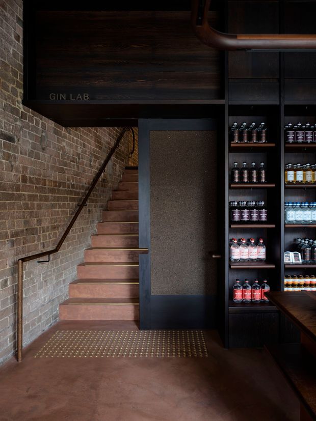 A moody commercial gin shop or tasting venue interior with an exposed brick stairwell, dark timber and black-painted built-in retail shelving stocked with bottles, and a stair landing with tactile brass studs. A narrow staircase leads upward beside a textured dark door, while a timber-clad overhead structure and copper-toned pipe detail reinforce the industrial character.