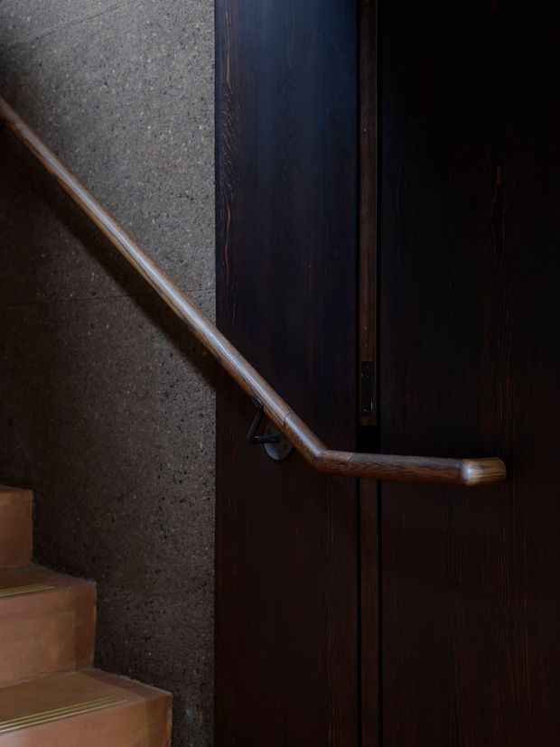 Close-up view of a stairway with a dark timber handrail and dark timber door or wall panelling beside a textured grey concrete wall. Warm terracotta-toned stair treads and subtle metal stair-edge strips are visible, creating a moody, tactile interior detail.