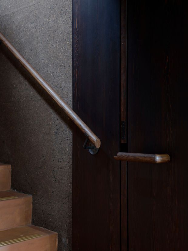 Close-up architectural detail of a dark timber stair and door area. A warm wood handrail runs along a rough grey concrete or masonry wall beside terracotta-toned stair treads, while a dark timber door with a matching horizontal handle occupies the right side. The image has a moody, tactile, material-focused character.